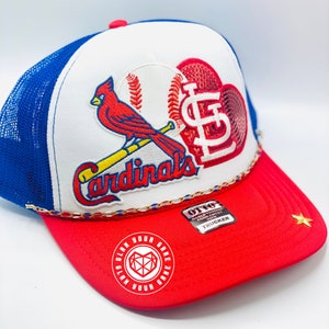 May include: A white, red, and blue trucker hat with a baseball-themed design. The hat features a red cardinal bird perched on a baseball bat, a baseball with a red heart, and the word "Cardinals" in gold lettering. The hat also has a gold chain trim and a red "Glam Your Game" logo on the side.