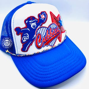 May include: A white and blue trucker hat with a red heart and star design. The heart has the word "Cubbies" in red lettering. The hat has a blue mesh back and a blue brim.