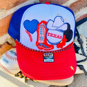 May include: A red, white, and blue trucker hat with a variety of embroidered patches, including a heart, a cowboy boot, a baseball, a cowboy hat, and a flower. The hat also has a gold chain trim and a label that reads "OTTO WORLD FAMOUS FOAM FRONT TRUCKER".