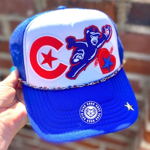 May include: Blue and white trucker hat with a red, white, and blue Chicago Cubs logo, a blue bear mascot, and a red heart with a blue star. The hat has a gold chain trim and a blue brim with the text "Glan Your Game Glan Your Game".