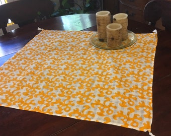 Puede incluir: Un camino de mesa blanco con un patrón floral amarillo y borlas en los extremos. Tres candelabros con un diseño de corteza de abedul están sobre un plato de vidrio en la parte superior del camino de mesa.