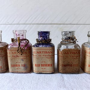 Könnte beinhalten: Fünf antike Glasflaschen mit Etiketten, jede mit einer einzigartigen Farbe und Textur. Die Etiketten tragen die Aufschrift "L'ARTISAN" und enthalten französischen Text. Die Flaschen sind in einer Reihe vor einem weißen Hintergrund angeordnet.