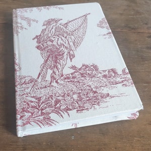 May include: A white notebook with a red and white toile pattern featuring a man with a fishing net. The notebook has a white spine and a red and white patterned cover.