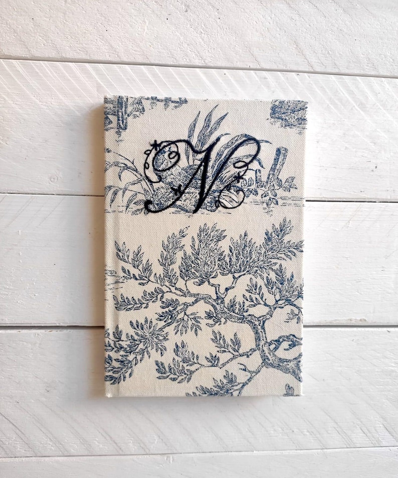 May include: A white fabric journal with a blue floral pattern and a black embroidered letter "N" on the front cover.