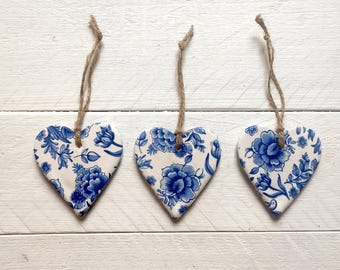 Op de afbeelding: Drie witte, hartvormige keramische ornamenten met blauwe bloemenpatronen en touwtjes om op te hangen.