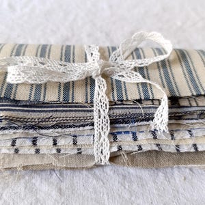 May include: A bundle of fabric remnants in shades of blue and cream, featuring striped and patterned designs. The fabrics are tied with a delicate white lace ribbon. The overall look is reminiscent of vintage textiles.