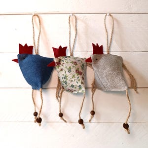 Op de afbeelding: Drie handgemaakte stoffen kippenversieringen die aan een touwtje hangen. De kippen zijn gemaakt van denim, bloemenstof en grijs stof. Elke kip heeft een rode kam en een rode keellap van vilt. De kippen hangen aan een wit houten oppervlak.