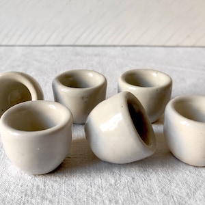 Könnte beinhalten: Sechs kleine, weiße Keramikbecher mit leicht strukturierter Oberfläche. Die Becher sind in einem Halbkreis auf einem weißen Stoffhintergrund angeordnet.