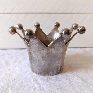 Könnte beinhalten: Ein silberfarbener, kronenförmiger Behälter mit strukturierter Oberfläche. Die Krone hat spitze Spitzen und kleine kugelförmige Verzierungen. Der Behälter wird wahrscheinlich zu Dekorationszwecken verwendet.