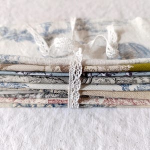 May include: A stack of fabric scraps in various shades of blue, brown, and white with a floral pattern. The fabric is tied together with a white lace ribbon.