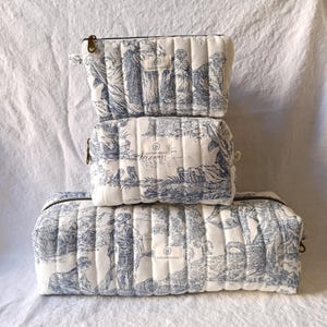 Peut inclure: Trois trousses de toilette matelassées empilées. Elles présentent un imprimé bleu et blanc de scène pastorale. Chaque trousse a une fermeture éclair et une petite étiquette en tissu. Les trousses sont de tailles différentes, la plus grande étant en bas.