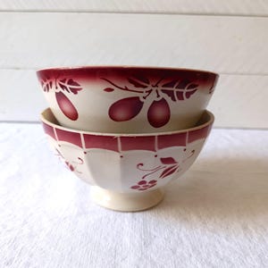 Pode incluir: Duas tigelas creme empilhadas. Elas têm uma borda vermelha e um desenho de frutas e folhas vermelhas. A tigela inferior tem uma base de pedestal. As tigelas estão em uma superfície branca.