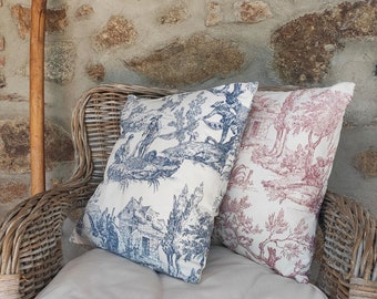 Puede incluir: Dos cojines cuadrados sobre una silla de mimbre. Un cojín presenta un patrón toile de jouy azul y blanco con escenas pastorales, mientras que el otro muestra un estampado toile de jouy rojo y blanco similar sobre un fondo claro.