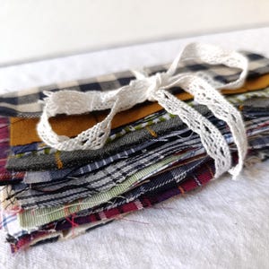 Puede incluir: Un montón de retales de tela en varios patrones y colores, incluyendo cuadros y tonos lisos. La tela está atada con una cinta de encaje blanco, creando un paquete ordenado. Las telas son en tonos de rojo, amarillo, verde y azul.