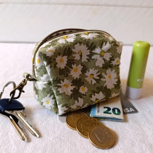 May include: A small, square, daisy-print fabric pouch with a zipper, holding a twenty-dollar bill, coins, a credit card, and a green lip balm. Keys are attached to the pouch with a metal clasp.