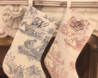 May include: Two personalized Christmas stockings with a toile pattern. The stocking on the left is blue and white and has the initial "M" embroidered on it. The stocking on the right is red and white and has the initial "L" embroidered on it.