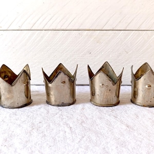 Puede incluir: Seis candeleros antiguos de metal plateado con forma de corona. Cada soporte tiene una base cilíndrica y un diseño de corona dentada y puntiaguda. Los candeleros están dispuestos en fila sobre una superficie blanca.