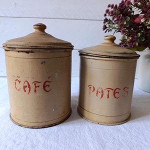 May include: Two antique beige canisters with lids. One canister has the word "CAFÉ" in red lettering, and the other has "PATES". The canisters are cylindrical and appear to be made of metal. The lids have a small knob on top.
