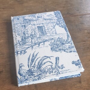 May include: A white notebook with a blue toile pattern featuring a house, a bridge, a woman, and a bird. The notebook has a white spine and blue edges.