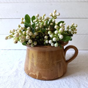 Könnte beinhalten: Eine braune Keramiktasse mit Henkel, gefüllt mit weißen Beerenstielen und grünen Blättern. Die Tasse hat ein rustikales, gealtertes Aussehen und steht auf einem weißen Tuch. Der Hintergrund ist eine weiße Holzwand.