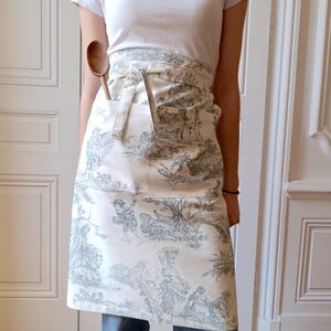 Op de afbeelding: Een witte schort met een blauw-wit toile-patroon. De schort heeft een grote zak aan de voorkant en wordt in de taille gebonden. Een houten lepel zit in de zak.