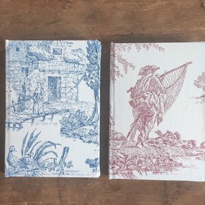 May include: Two small, white, fabric-covered journals with a vintage-style design. The journal on the left has a blue design of a house and trees. The journal on the right has a red design of a person with a fishing net.