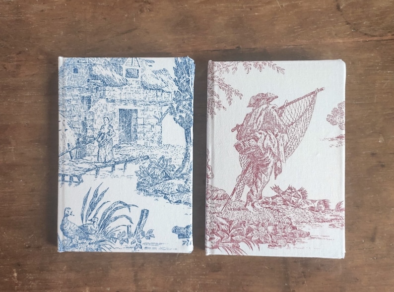 May include: Two small, white, fabric-covered journals with a vintage-style design. The journal on the left has a blue design of a house and trees. The journal on the right has a red design of a person with a fishing net.