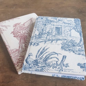 May include: Two fabric-covered journals with a vintage toile pattern. One journal has a red and white design with a bird and floral motif. The other journal has a blue and white design with a bird and floral motif.