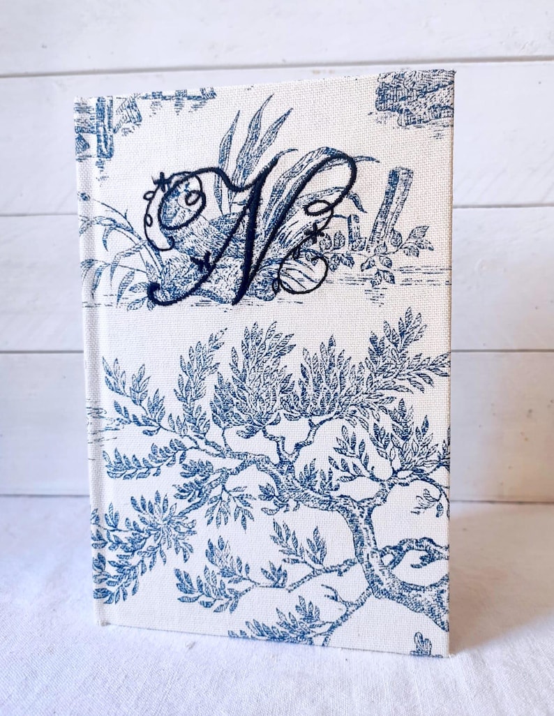 May include: A white linen journal with a blue and white toile pattern. The journal has a black embroidered letter "N" on the front cover.