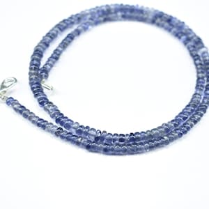 K&ouml;nnte beinhalten: Eine Halskette aus kleinen, runden, blau-violetten Perlen. Die Perlen sind zu einer zarten Kette aufgereiht. Ein silberfarbener Verschluss ist an einem Ende befestigt. Die Halskette wird vor einem wei&szlig;en Hintergrund gezeigt.