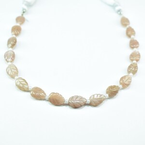May include: A necklace featuring carved leaf-shaped beads in a warm, peach color. The beads are strung together with small, white beads, creating a delicate and natural design. The necklace is displayed against a white background.