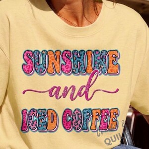 以下が含まれることがあります： 「SUNSHINE and ICED COFFEE」という文字がプリントされたペールイエローの半袖Tシャツ。文字はピンク、オレンジ、ブルーの色合いのカラフルなヒョウ柄で塗りつぶされており、暗い色の縁取りが施されています。