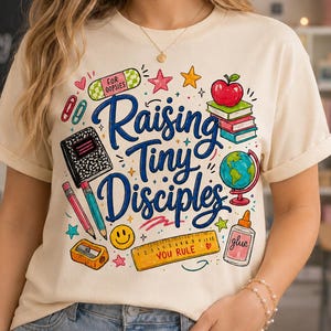 May include: A cream-colored t-shirt featuring a colorful graphic print with the text "Raising Tiny Disciples" in blue script. The design includes school supplies like a notebook, pencils, a globe, a ruler labeled "YOU RULE", and an apple.
