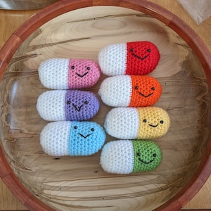 May include: Seven crocheted pill-shaped plushies in a wooden bowl. The plushies are white with a different colored half, each with a smiling face. The colors are red, pink, purple, blue, yellow, green, and orange.