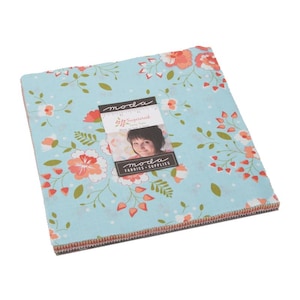 May include: A stack of fabric squares with a floral pattern in shades of light blue, coral, and green. The fabric features a repeating design of flowers and leaves. A label with the text "moda FABRICS + SUPPLIES" is attached to the fabric.