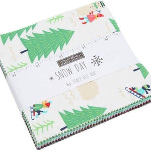 May include: A stack of square fabric pieces featuring a white background with green pine trees and figures sledding. A white paper band wraps around the center, printed with "SNOW DAY by STACY IEST HSU" and a ruler measuring up to 8 inches.