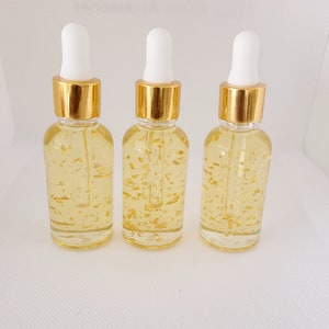 K&ouml;nnte beinhalten: Drei transparente Glas-Tropfflaschen, gef&uuml;llt mit einer gelben Fl&uuml;ssigkeit und Goldflocken. Jede Flasche hat einen goldenen Deckel und eine wei&szlig;e Pipette. Die Flaschen sind auf wei&szlig;em Hintergrund in einer Reihe angeordnet.