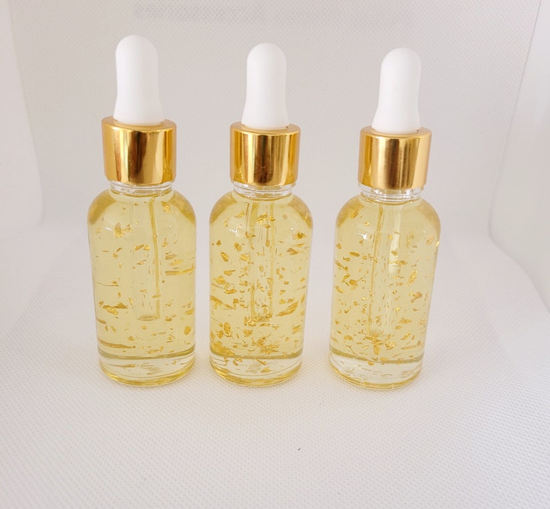 K&ouml;nnte beinhalten: Drei transparente Glas-Tropfflaschen, gef&uuml;llt mit einer gelben Fl&uuml;ssigkeit und Goldflocken. Jede Flasche hat einen goldenen Deckel und eine wei&szlig;e Pipette. Die Flaschen sind auf wei&szlig;em Hintergrund in einer Reihe angeordnet.