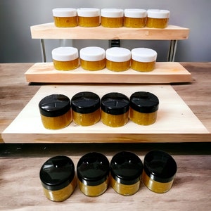 May include: A set of 11 small glass jars with black lids, filled with a yellow substance. The jars are arranged on a two-tiered wooden display stand.