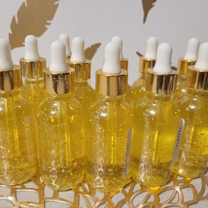 K&ouml;nnte beinhalten: Eine Sammlung klarer Glasflaschen, gef&uuml;llt mit einer goldenen Fl&uuml;ssigkeit und goldenen Flocken. Jede Flasche hat eine wei&szlig;e Pipette und einen goldfarbenen Deckel. Die Flaschen stehen auf einer goldfarbenen dekorativen Oberfl&auml;che.