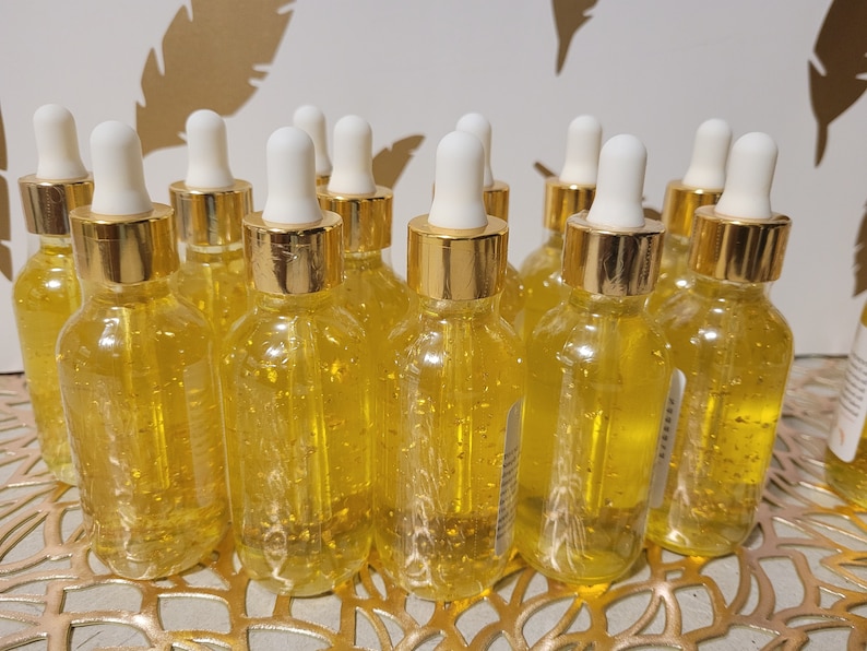 K&ouml;nnte beinhalten: Eine Sammlung klarer Glasflaschen, gef&uuml;llt mit einer goldenen Fl&uuml;ssigkeit und goldenen Flocken. Jede Flasche hat eine wei&szlig;e Pipette und einen goldfarbenen Deckel. Die Flaschen stehen auf einer goldfarbenen dekorativen Oberfl&auml;che.