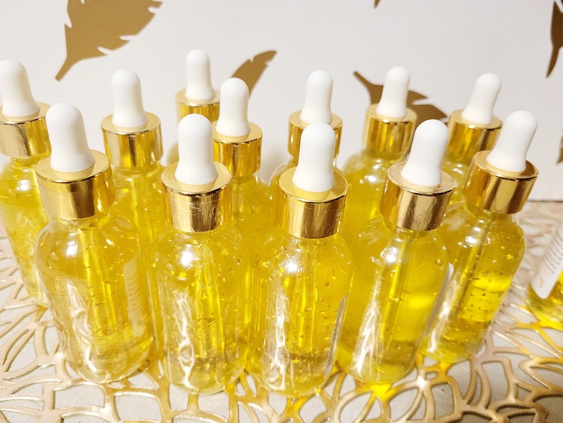 K&ouml;nnte beinhalten: Eine Ansammlung klarer Glasflaschen, gef&uuml;llt mit einer goldenen Fl&uuml;ssigkeit. Jede Flasche hat eine wei&szlig;e Pipette und einen goldfarbenen Verschluss. Die Flaschen stehen auf einer dekorativen, goldenen Oberfl&auml;che, vermutlich ein Hautpflege- oder Sch&ouml;nheitsprodukt.