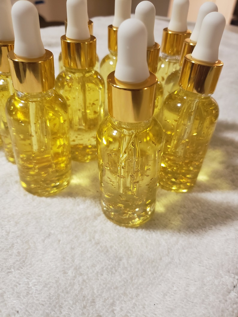 K&ouml;nnte beinhalten: Eine Sammlung kleiner Glasflaschen, gef&uuml;llt mit einer gelben Fl&uuml;ssigkeit und Goldflocken. Jede Flasche hat einen goldfarbenen Verschluss und eine wei&szlig;e Pipette. Die Flaschen sind auf einer wei&szlig;en, strukturierten Oberfl&auml;che angeordnet.
