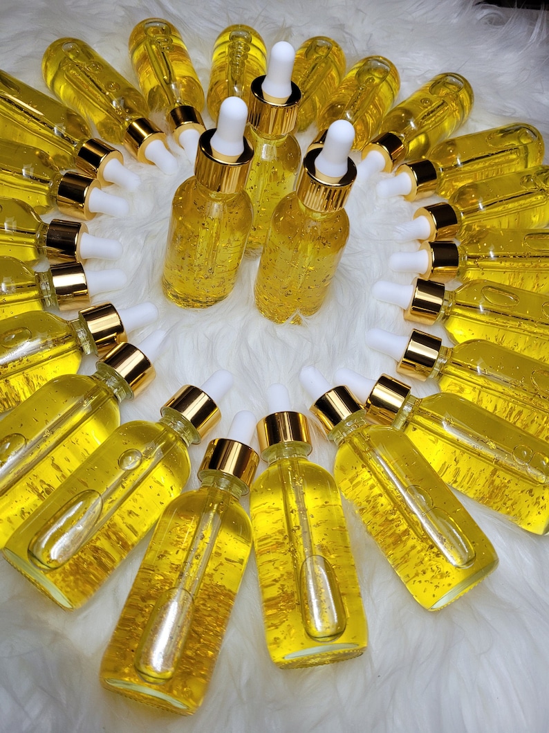 K&ouml;nnte beinhalten: Eine Anordnung kleiner, klarer Glasflaschen, gef&uuml;llt mit einer goldenen Fl&uuml;ssigkeit und Goldflocken. Jede Flasche hat eine wei&szlig;e Pipette und einen goldfarbenen Deckel. Die Flaschen sind kreisf&ouml;rmig auf einer wei&szlig;en, flauschigen Oberfl&auml;che angeordnet.