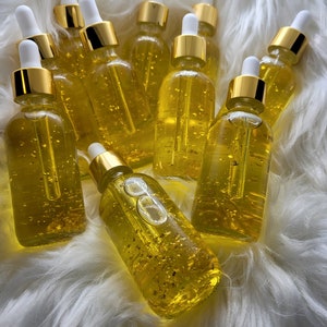 K&ouml;nnte beinhalten: Eine Sammlung klarer Glasflaschen, gef&uuml;llt mit einer goldenen Fl&uuml;ssigkeit und Goldflocken. Jede Flasche hat eine wei&szlig;e Pipette und einen goldenen Verschluss. Die Flaschen sind auf einer flauschigen, wei&szlig;en Oberfl&auml;che angeordnet.