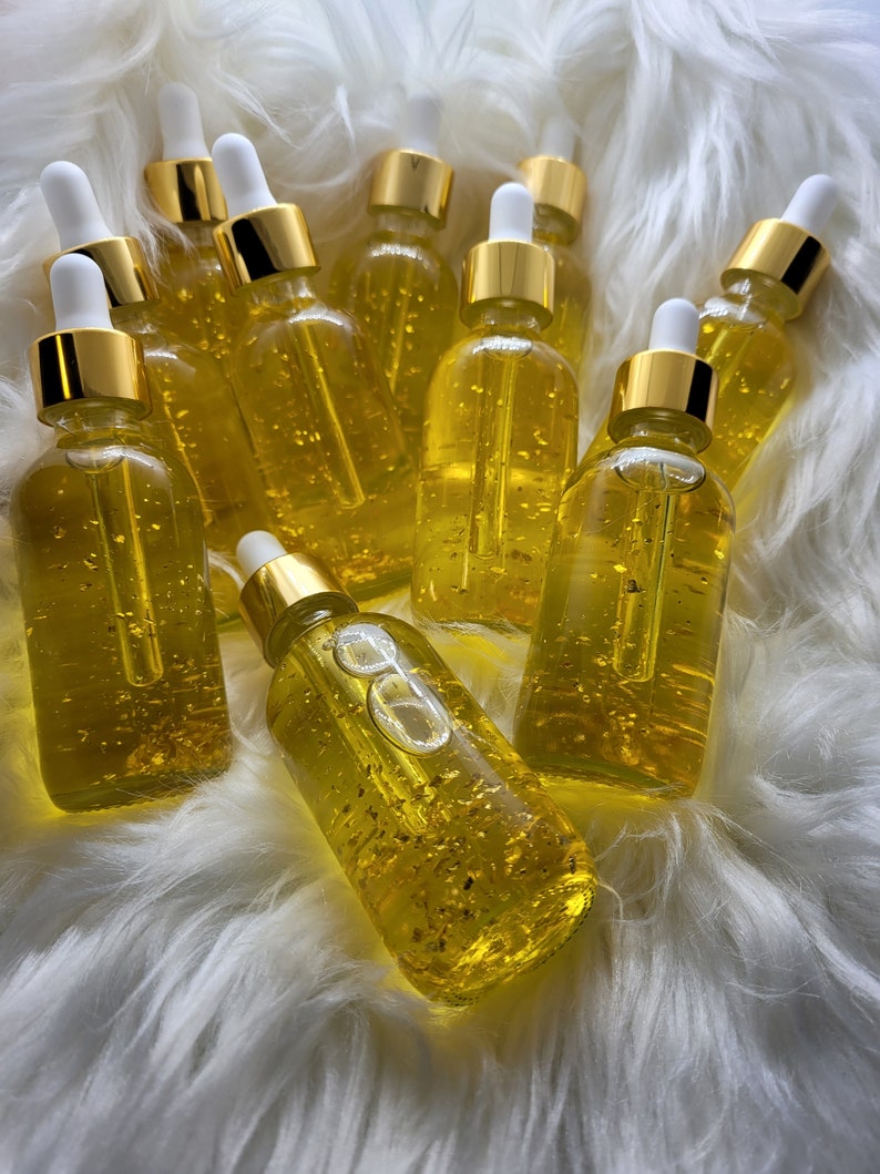 K&ouml;nnte beinhalten: Eine Sammlung klarer Glasflaschen, gef&uuml;llt mit einer goldenen Fl&uuml;ssigkeit und Goldflocken. Jede Flasche hat eine wei&szlig;e Pipette und einen goldenen Verschluss. Die Flaschen sind auf einer flauschigen, wei&szlig;en Oberfl&auml;che angeordnet.