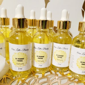 K&ouml;nnte beinhalten: Eine Sammlung klarer Glasflaschen, gef&uuml;llt mit einer goldenen Fl&uuml;ssigkeit, jede mit einem wei&szlig;en Etikett mit der Aufschrift "24 KARAT FACIAL Serum" und "2 OZ". Die Flaschen haben goldfarbene Kappen und wei&szlig;e Tropfer. Der Markenname "Onu Latte's Beauty" ist ebenfalls sichtbar.