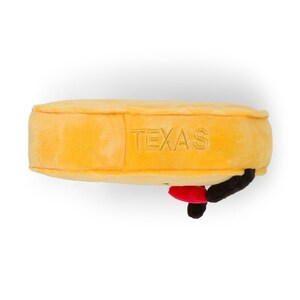 Puede incluir: Un objeto rectangular de felpa amarilla con la palabra "TEXAS" bordada en el lateral. Tiene un acento rojo y negro. Este art&iacute;culo es probablemente una novedad o un recuerdo.