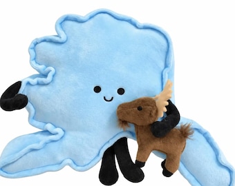 May include: A light blue plush toy shaped like the state of Alaska with a smiling face, black limbs, and a small brown plush moose attached to its front. The Alaska-shaped toy features a soft texture and simple embroidered facial features.