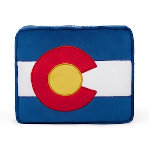 Puede incluir: Una almohada de felpa azul, blanca y roja con forma de la bandera del estado de Colorado. La bandera presenta un círculo rojo grande con un círculo amarillo en su interior y la letra "C" en el centro.